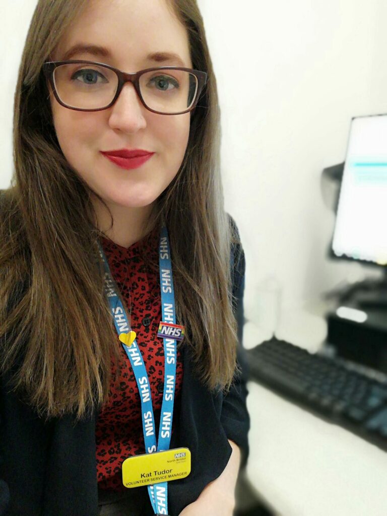 Volunteer Services Manager Kat Tudor sits at her desk. She is wearing a red top, black blazer and blue lanyard.
