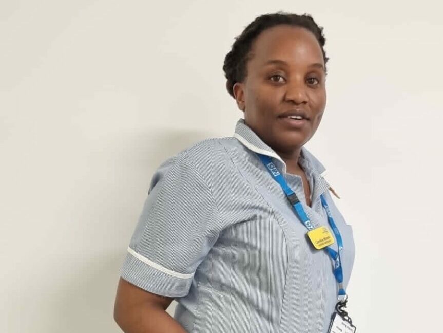 Nurse Associate Caroline is pictured standing in front of a while wall. She is wearing her uniform of a striped top and navy trousers..