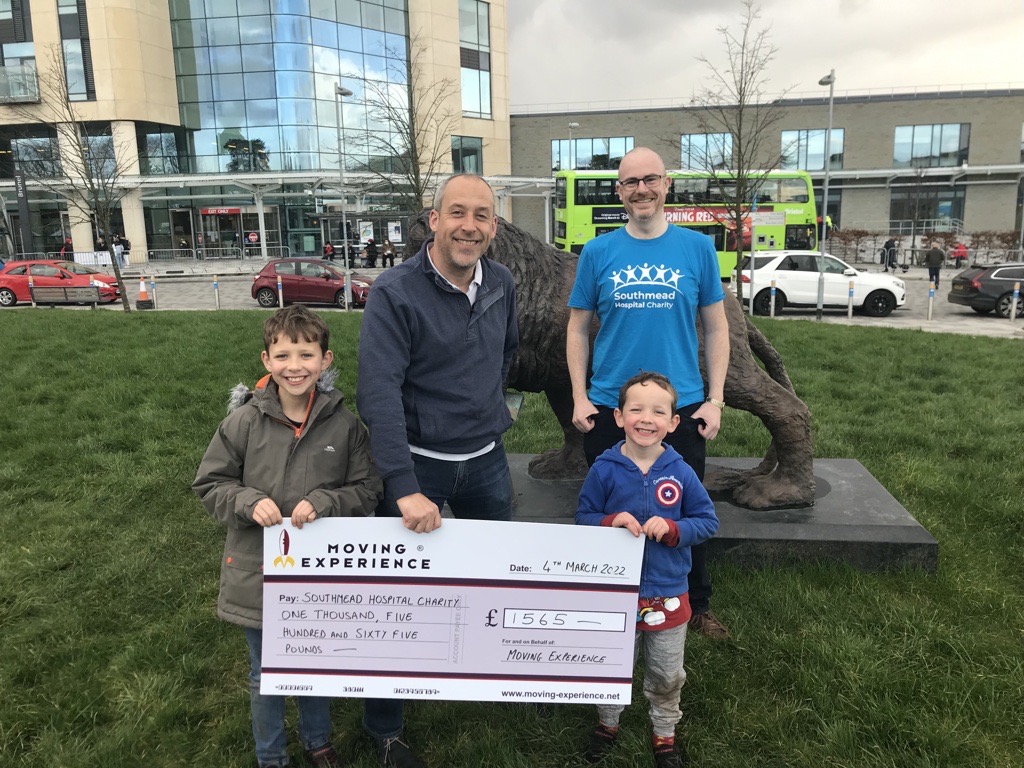 Fundraiser Adrian stands with Matt from Moving Excellence and Matt's two sons, all holding a novelty sized cheque